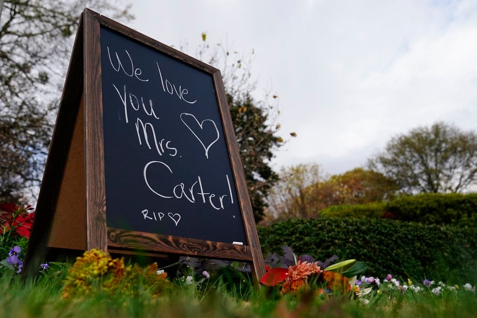 Bouquets of flowers and a chalkboard sign that reads "We love you Mrs. Carter" is displayed...